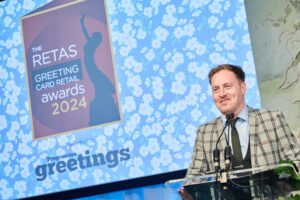 An awards ceremony with a graphic logo on a big screen stating the Reta awards. A man with short brown hair and wearing a brown and beige check jacket, smiles as he addresses the audience, standing at a glass console and speaking into a microphone.