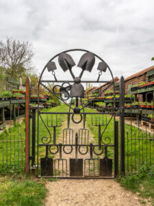 A closed metal gate formed out of spade and shovel heads. The gate leads to a gravel path flanked by grass verges and stands full of a variety of garden plants.